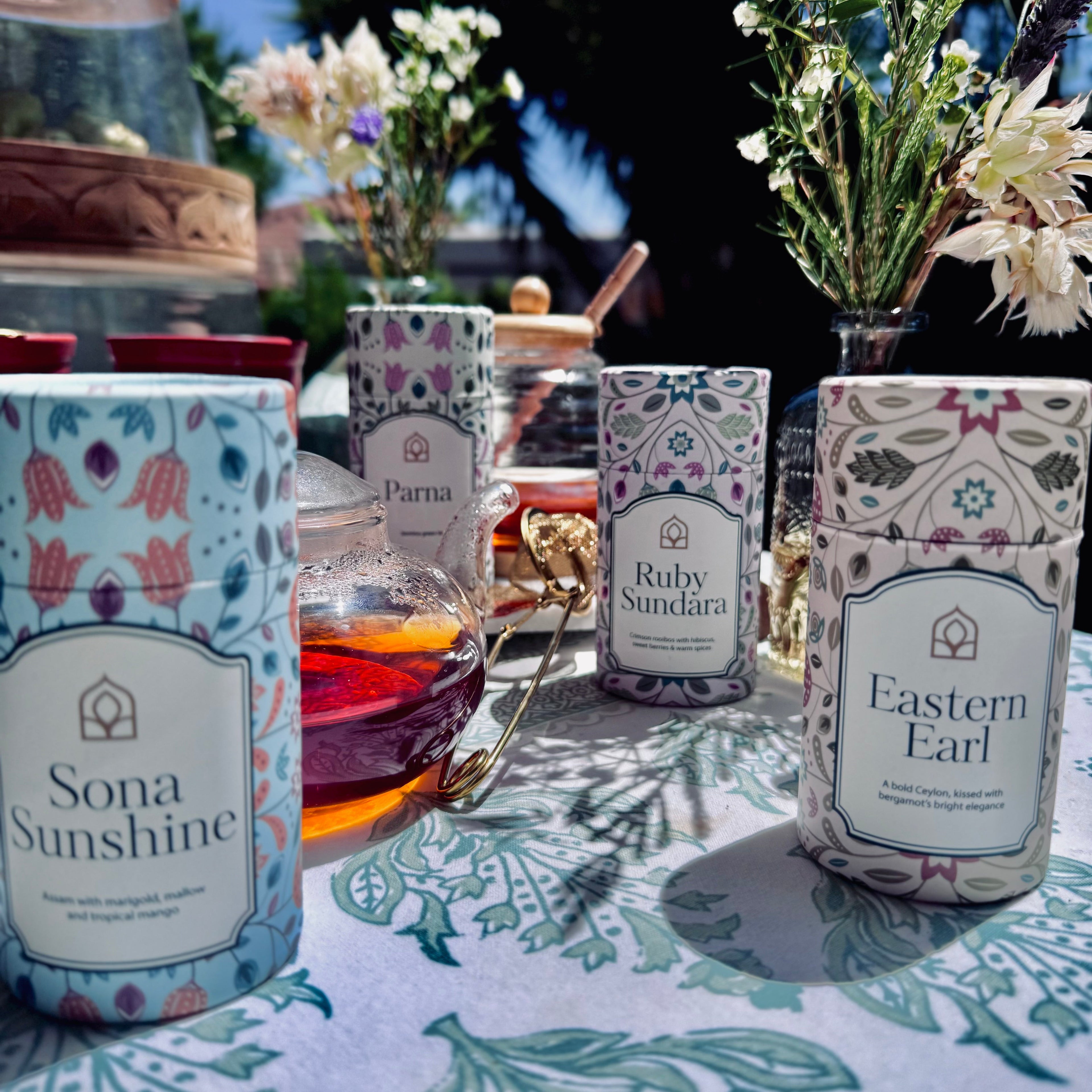 Chai's four premium loose leaf tea canisters with a glass cup of brewed tea, styled outdoors with flowers and natural sunlight