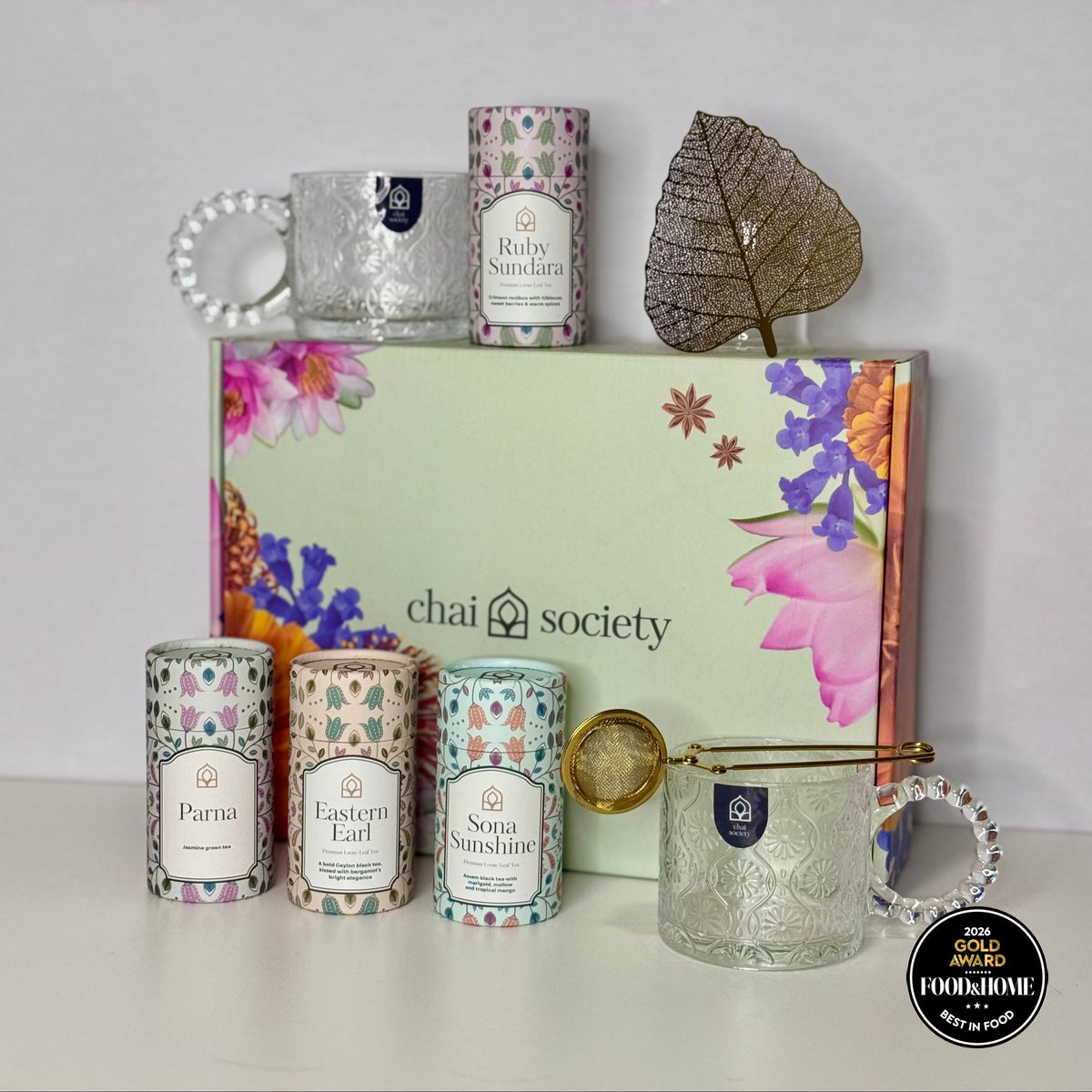 A hand-crafted Chai Society tea gift box opened to show reusable glass teacup, infuser and gold teaspoon, presented gift-ready with no wrapping required