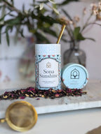 Tea canister labeled 'Sona Sunshine' with a gold spoon on a marble surface with dried tea leaves. As well as gold tea strainer.